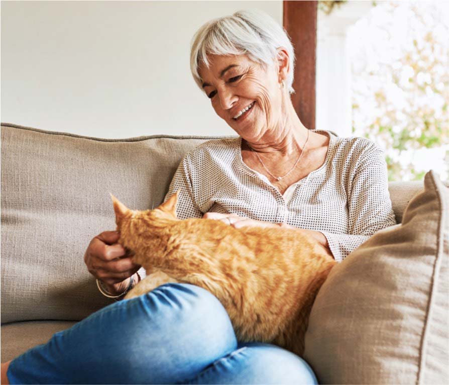 Elderly woman joyfully pets her orange cat while sitting on a comfortable beige sofa at home.