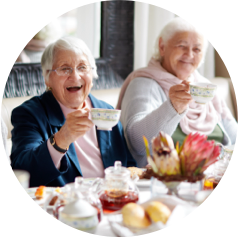 Two elderly women enjoying tea and pastries at a cozy table, smiling and raising their teacups.