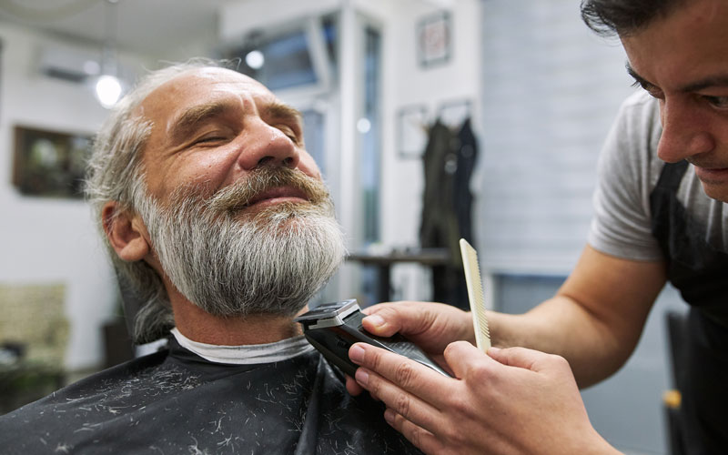 A barber trimming an older mans beard while he relaxes in a modern salon.
