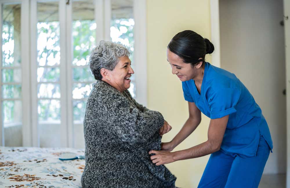 Caregiver in blue scrubs assisting an elderly woman with a smile in a cozy home setting.