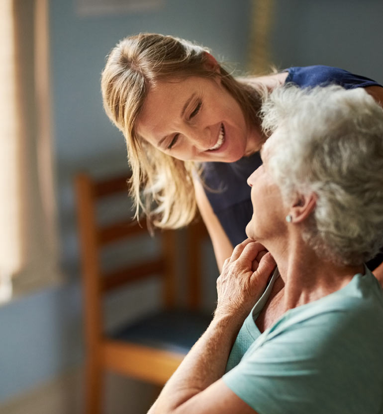 Caregiver smiling at and assisting an elderly woman in a memory care facility.
