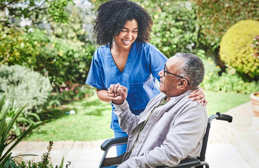 Caregiver in blue scrubs smiling and assisting senior man in a wheelchair in a garden setting.