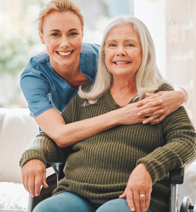 A senior woman in a wheelchair smiling with a caregiver embracing her from behind in a cozy setting.