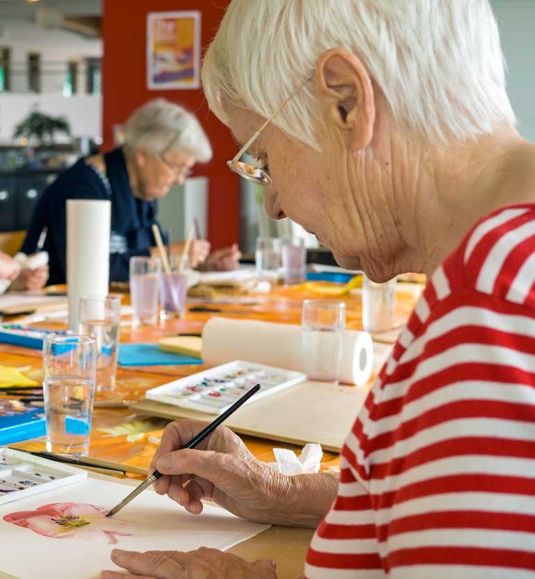 Elderly woman painting with watercolors in an art class alongside other seniors.