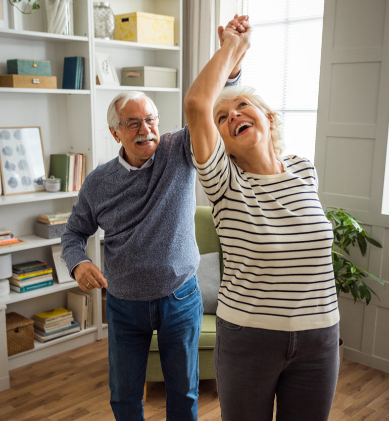 Elderly couple joyfully dancing together in a cozy living room with bookshelves in the background.