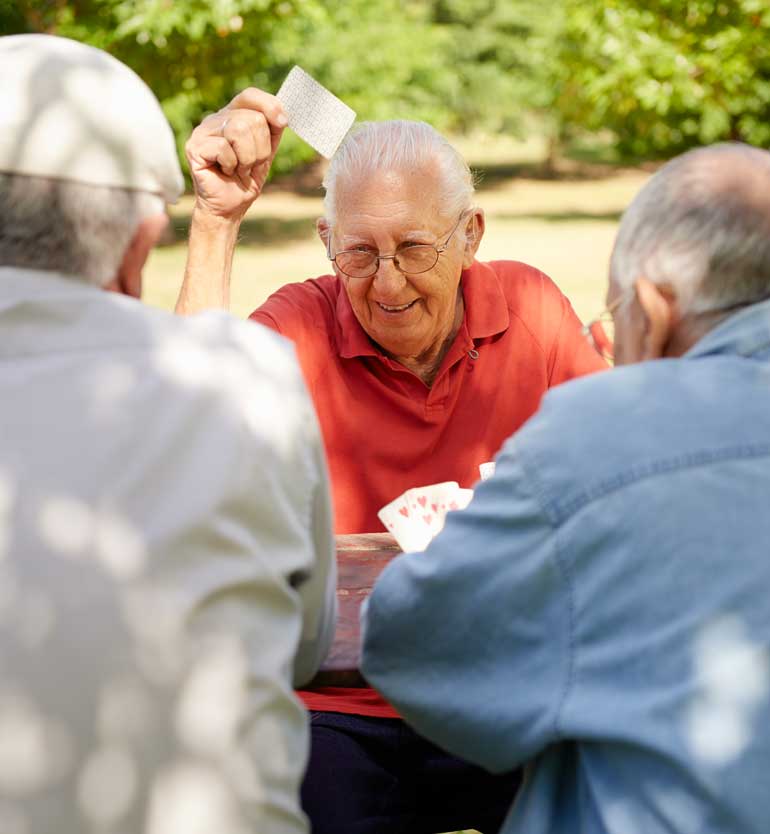 Three elderly men enjoying a card game outdoors, with one holding up a card and smiling.