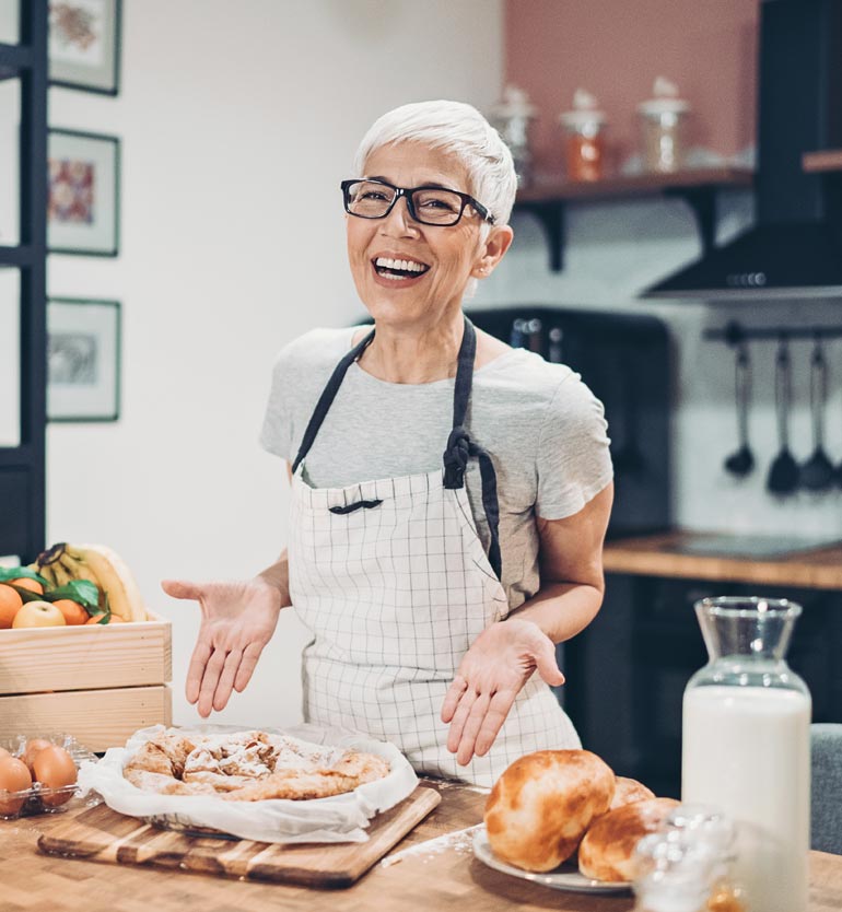 A smiling elderly woman wearing glasses and an apron showcases her freshly baked pastries in the kitchen.
