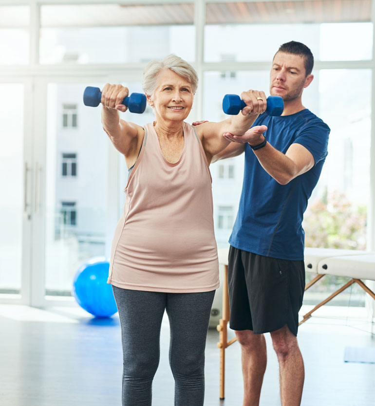 Elderly woman lifting dumbbells with assistance of a trainer in a bright fitness gym.