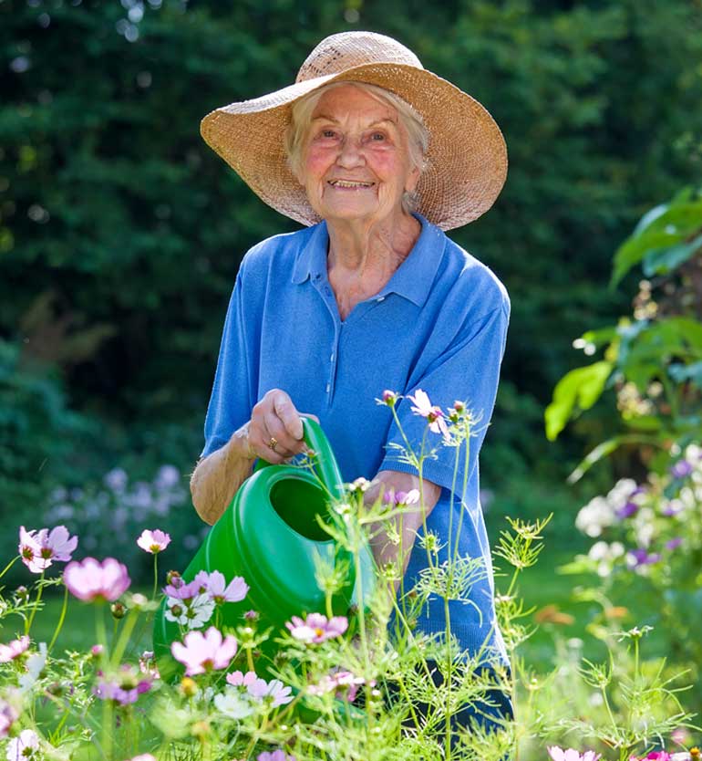An elderly woman watering flowers in a garden, wearing a blue shirt and a large straw hat.