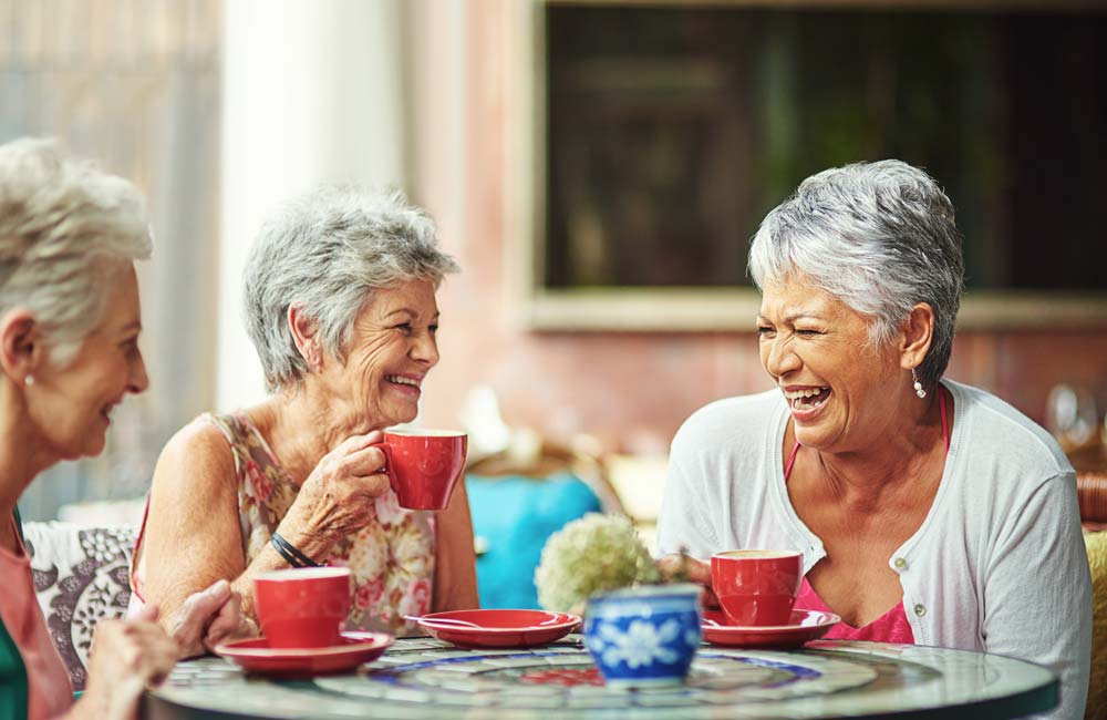 Three elderly women enjoy coffee and laugh together at an outdoor table with red cups and saucers.