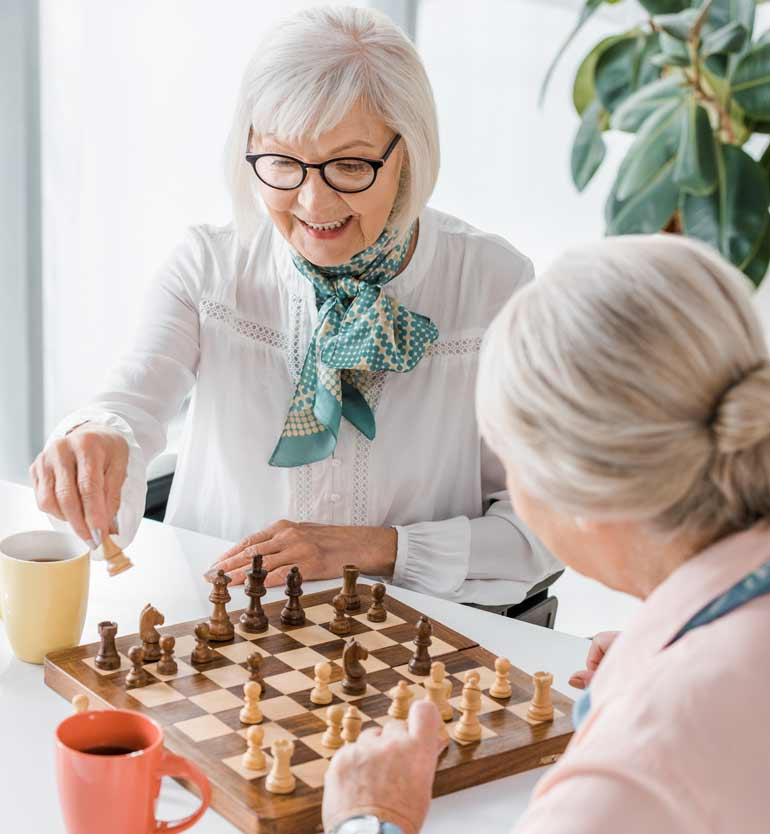 Two elderly women enjoying a game of chess at a bright, modern space with coffee mugs on the table.