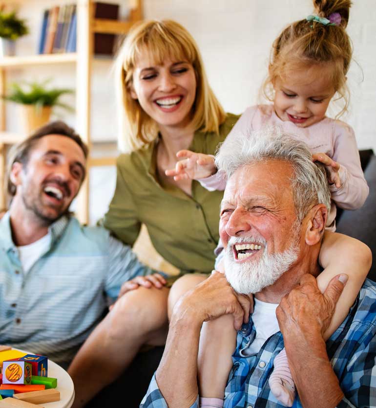 Senior man with white beard laughing with family, including a child on his shoulders, in a cozy setting.