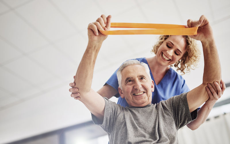 A senior man smiling while lifting an exercise band under the guidance of a female nurse.