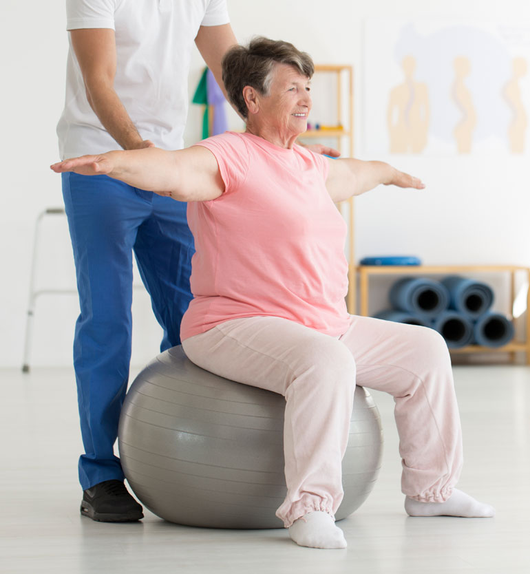 Senior woman doing arm exercises on a stability ball with therapist in a bright, airy room.