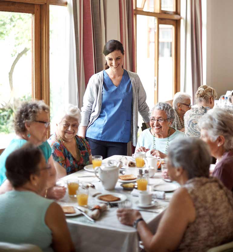 Senior women sharing a meal and smiling with a caregiver in the dining area of an assisted living facility.