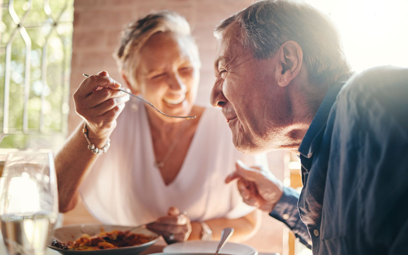 Elderly woman playfully feeding an elderly man with a spoon at an outdoor dining table.
