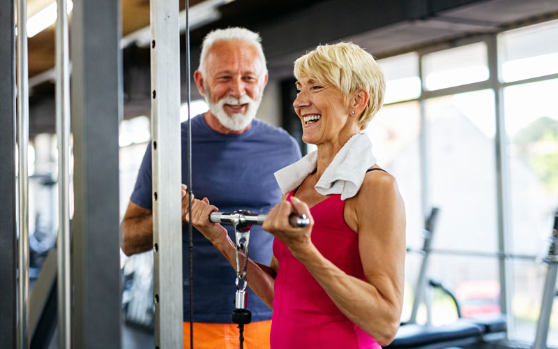 Seniors exercising in a gym, woman using a machine while smiling, man observing and encouraging.