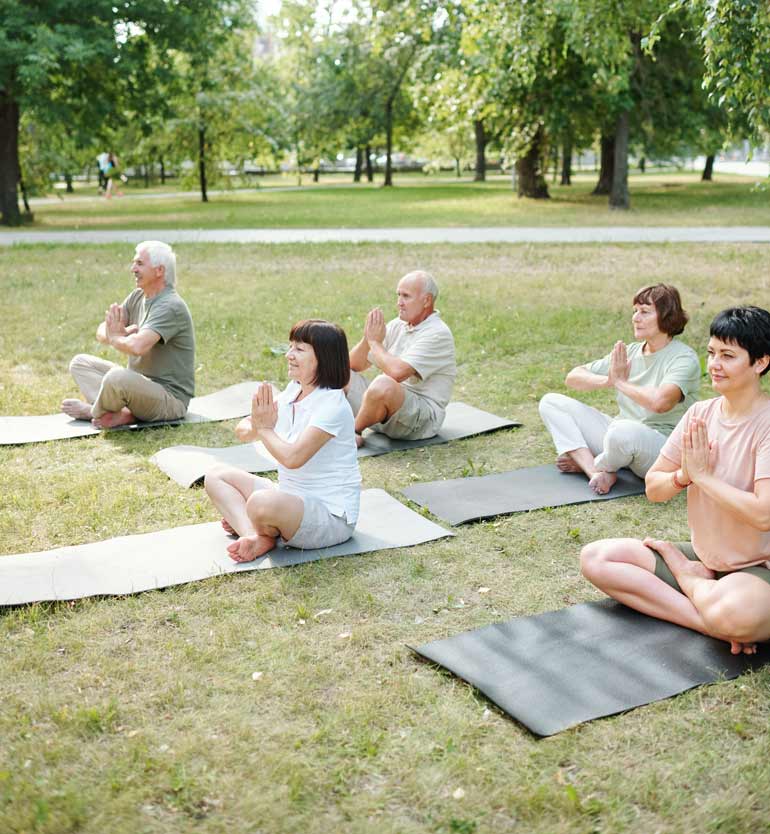 Seniors practicing yoga outdoors on mats in a park, seated with hands in prayer position