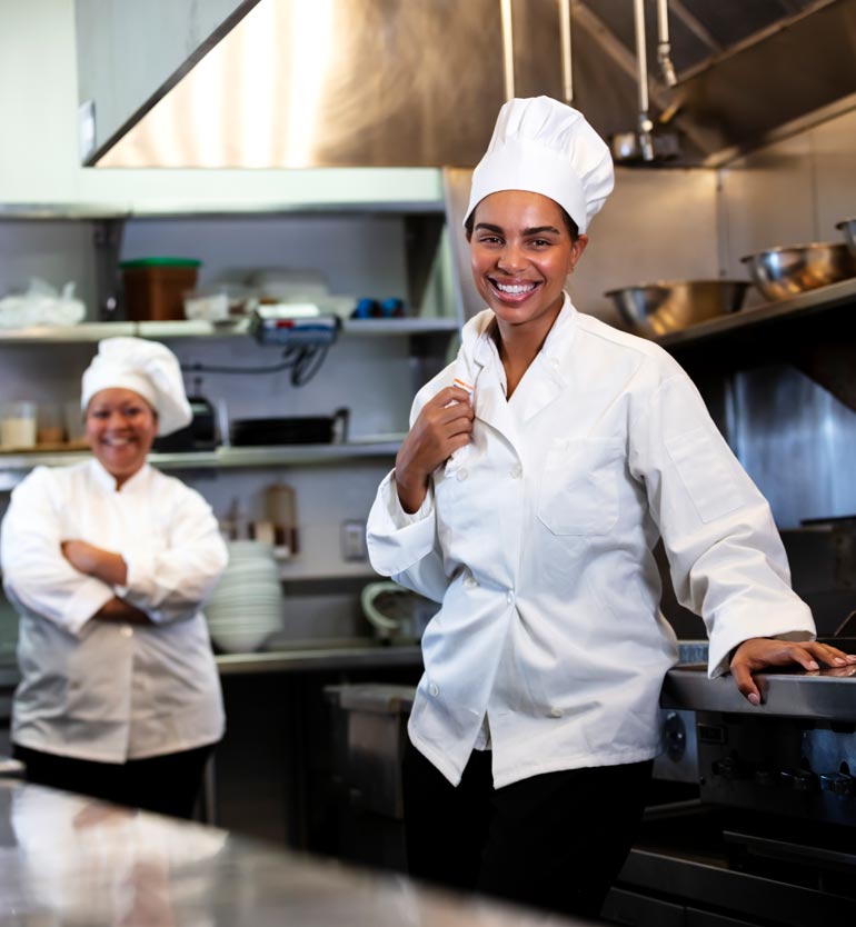 Two smiling chefs in a commercial kitchen, one in the foreground and one in the background